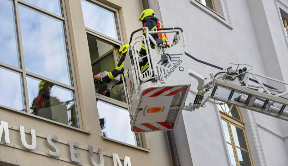 Feuerwehrmänner nehmen auf ihrer Drehleiter oben an der Fassade etwas aus einem offenen Fenster entgegen.