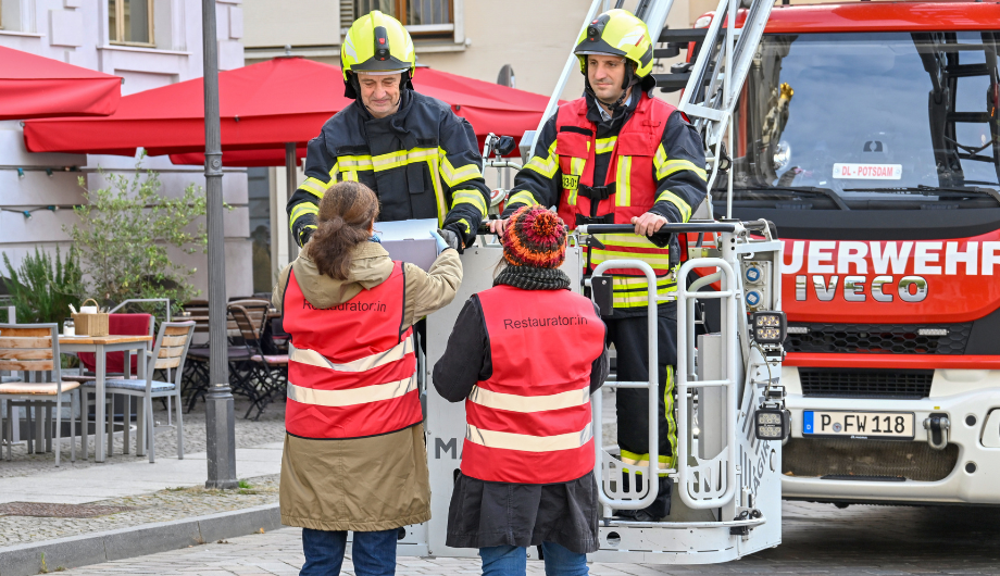 Restauratorinnen vor dem Museum nehmen den Gegenstand den Feuerwehrmännern entgegen.