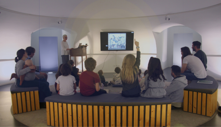 Eine Gruppe Kinder sitzt auf Sitzbänken in einem Raum und schaut auf einen Bildschirm.