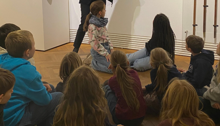 Eine gruppe Kinder sitzt auf dem Boden auf Matten und hört einem Vortrag zu.